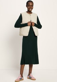 Helle beige gepolsterte Weste über einem dunkelgrünen gerippten Kleid, kombiniert mit rosa Sneakers mit blauen Akzenten. Neutraler Hintergrund.