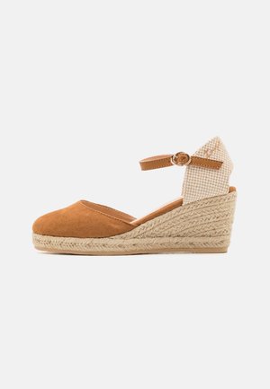 Zapatos de cuña de ante marrón con una plataforma de yute estilo espadrille. Presentan una correa alrededor del tobillo de color beige tejida con un detalle de hebilla.