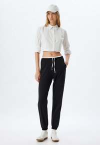 Witte cropped button-up shirt, zwarte sweatpants met een elastische tailleband en witte sneakers. De pet is lichtgrijs.