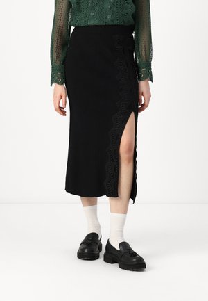 Falda negra hasta la rodilla con una abertura lateral, que presenta intrincados detalles de encaje en el lado izquierdo. Llevada con zapatos negros y calcetines blancos.