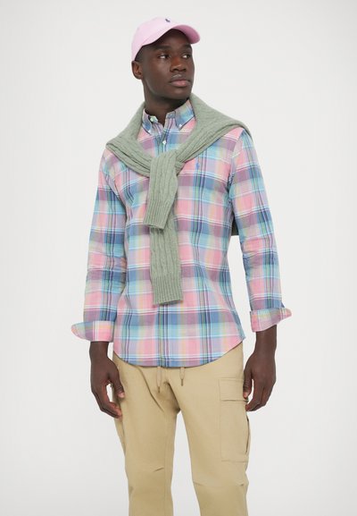 Večbarvna karo srajca v modro-roza-zelenih tonih; svetlo zeleni pleteni pulover, premetan čez ramena; svetlo bež hlace s cargo žepi; roza kapa.