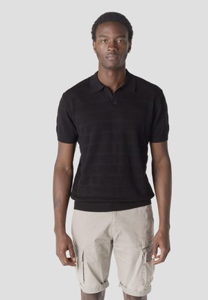 Fiatal férfi fekete rövid ujjú kötött pólóban és bézs színű tereptárgy rövidnadrágban, sima világosszürke háttér előtt áll.