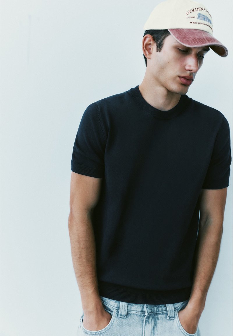 Jeune homme portant une chemise à manches courtes sombre, un jean denim clair et une casquette de baseball blanche et rouge, debout les mains dans les poches.