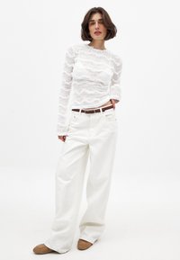 Top bianco in pizzo con maniche lunghe e motivo a onde, abbinato a jeans bianchi a vita alta a gamba larga e scarpe basse marroni.