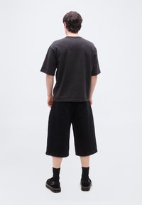 Άνδρας που φοράει μαύρο υπερμεγέθες T-shirt, μαύρες βερμούδες μέχρι το γόνατο, μαύρες κάλτσες και μαύρα σανδάλια, στέκεται με πλάτη προς την κάμερα πάνω σε λευκό φόντο.