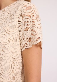 Beżowa koronkowa bluzka z falbaniastymi rękawami, misternymi wzorami kwiatowymi i półprzezroczystą teksturą, podkreślająca delikatne rzemiosło.