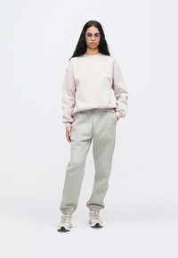 Γυναίκα με μακριά καστανά μαλλιά που φοράει ελαφρώς μωβ φούτερ, γκρίζες φόρμες, γυαλιά ηλίου με μωβ απόχρωση και λευκά sneakers σε λευκό φόντο.