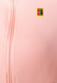 Chaqueta rosa claro con textura suave, cremallera frontal y logo multicolor bordado en el lado izquierdo.