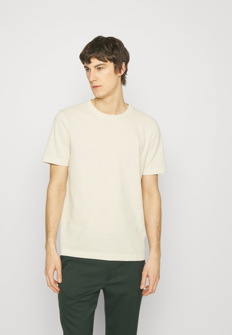 Lys beige kortærmet t-shirt med rund halsudskæring, glat tekstur og en afslappet pasform, kombineret med mørkegrønne bukser.