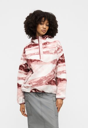 Femme aux cheveux bouclés portant un pull polaire rose et blanc à motifs et une jupe grise, debout devant un fond uni.