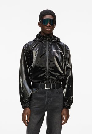 Homme portant une veste zippée noire brillante, des lunettes de soleil noires, une ceinture noire et un jean gris foncé, debout devant un fond blanc.