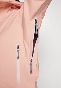 Světle růžová nepromokavá bunda s šedými zipy a nastavitelného stahovacími prvky. Texturovaná tkanina s úhlovými zipovými kapsami po stranách.