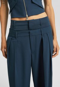 Marinblå skräddarsydda byxor med hög midja, veckad fram, och sidofickor, tillverkade av ett slätt, strukturerat tyg. Dragkedja som stängning.