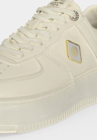 Κοντινή λήψη ενός κρεμ sneaker με κορδόνια, υφασμένη σόλα και τετράγωνο λογότυπο με χρυσή επένδυση στο πλάι.