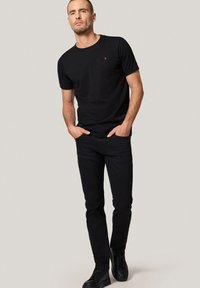 Mężczyzna z krótkimi włosami, ubrany w czarną koszulkę, czarne dżinsy i czarne trampki, stojący z rękami w kieszeniach na jednolitym tle.