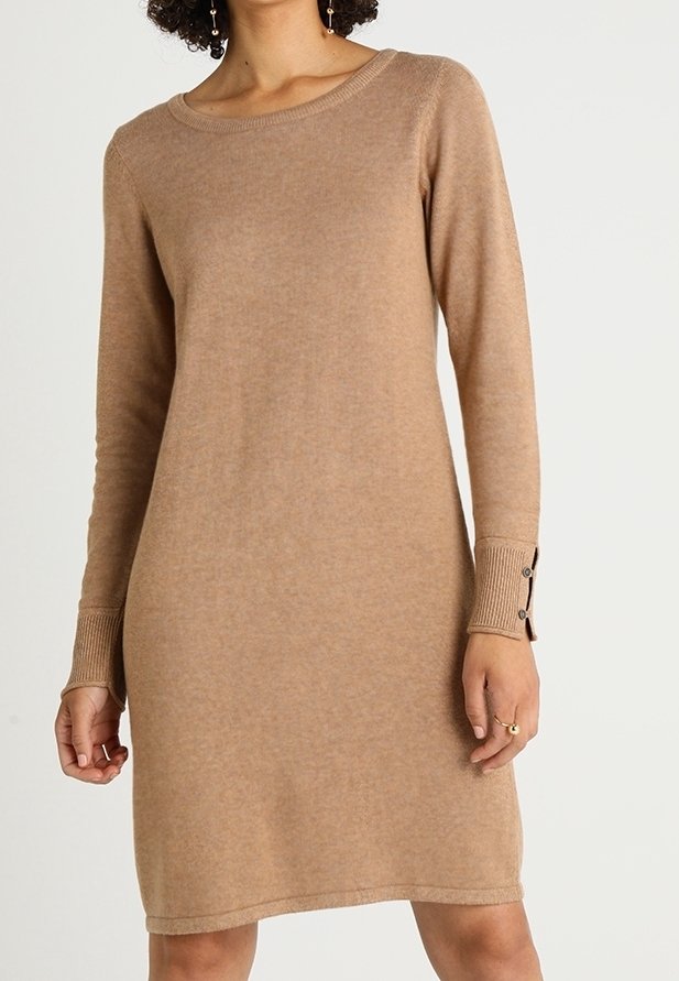 Femme portant une robe tricotée couleur camel à manches longues avec des détails de boutons aux poignets, debout devant un fond uni.