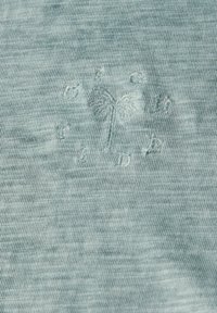Tela azul claro con una textura sutil, que presenta un diseño bordado de una palmera y las iniciales "D". Acentos de letras espaciadas alrededor del diseño.
