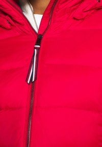 Veste rembourrée rouge avec fermeture éclair, dotée d'une tirette en ruban rayé. Texture lisse avec des panneaux matelassés et un col haut.