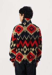 Red, black, and beige patterned fleece pullover with geometric designs, high collar, and elastic cuffs. Soft texture, relaxed fit.