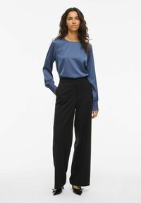 Vrouw met krullend haar draagt een blauwe satijnen blouse met lange mouwen en smockmanchetten, zwarte wijde broek en zwarte puntige hakken.