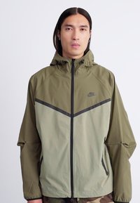 Ung mand med langt mørkt hår iført en olivenfarvet og lys grøn Nike vindjakke med sort lynlås, stående mod en ensfarvet baggrund.
