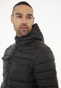 Chaqueta de plumas negra con capucha, con textura acolchada, cierre frontal con cremallera y dos bolsillos de parche. Logo en relieve en la manga.