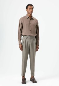 Bruin overhemd met lange mouwen en knoopsluiting, gecombineerd met lichtgrijze geplooide broek en donkerbruine loafers. Glad weefsel overal.