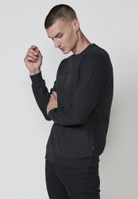 Sudadera negra texturizada con cuello redondo y puños acanalados, que presenta un panel frontal liso y una silueta ajustada.