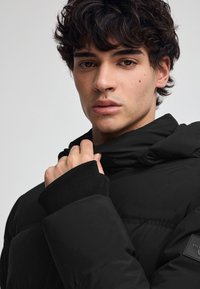 Svart pufferjacka med högt krage, ribbade ärmslut och en mjuk textur. Har en patch-logotyp på ärmen och en slät, glänsande yta.