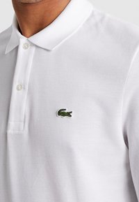 Vit polo skjorta i texturerat tyg, med en tre-knapps knäppning och en liten grön krokodil-logotyp på vänster bröst.