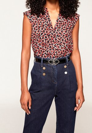 Kobieta w czarnej koszuli bez rękawów z czerwonymi i białymi wzorami serc, włożonej do ciemnoniebieskich dżinsów z wysokim stanem, ozdobionych metalowymi guzikami i czarnym paskiem.