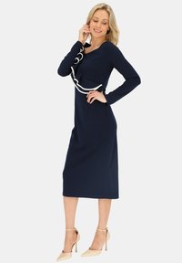 Granatowa sukienka z długim rękawem o dopasowanym kroju, z płynącym białym akcentem wzdłuż dekoltu i długości do kolan. Beżowe szpilki.
