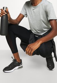 Nike grå t-shirt med kort ärm, svarta leggings med mesh-sektioner och svarta sneakers med vita detaljer. Håller i en svart isolerad vattenflaska.