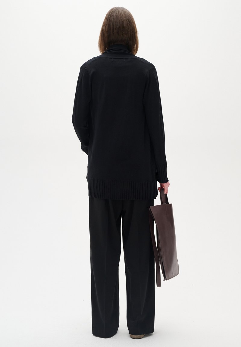 Schwarze langärmelige Strickjacke mit geripptem Saum, kombiniert mit lockeren schwarzen Hosen. Das Model hält eine dunkelbraune Leder-Totebag.