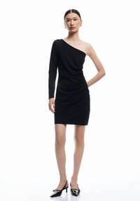 Schwarzes, einseitig-trägendes Kleid mit gerafften Details, tailliertem Schnitt und langen Ärmeln. Getragen mit schwarzen Lackschuhen. Glatte Textur.