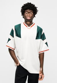 Λευκό αμάνικο πλεκτό με πράσινες και κόκκινες λεπτομέρειες, με κοντά μανίκια, V-λαιμόκοψη και άνετη εφαρμογή. Λογότυπο Adidas στο στήθος.