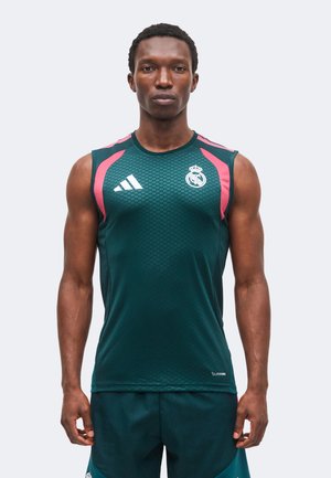 Uomo atletico che indossa una canotta sportiva verde scuro con accenti rosa e i loghi del Real Madrid e Adidas, in piedi su uno sfondo semplice.