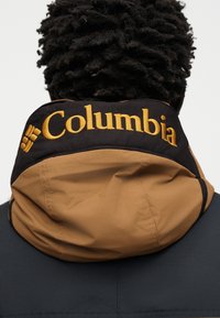 Personne aux cheveux bouclés portant une veste noire et marron avec un col relevé brodé du mot « Columbia » en jaune.