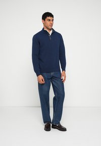 Camisola de malha azul-marinho com gola de fecho de correr e punhos canelados, combinada com jeans de ganga azul e sapatos castanho-escuros, em pé contra um fundo simples.