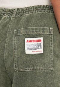 Zaļie džinsu augstās jostas bikses ir aprīkotas ar savilkta elastīga vidukļa joslu, aizmugures kabatu un reklamējošu etiķeti ar tekstu sarkanā un zilā krāsā.