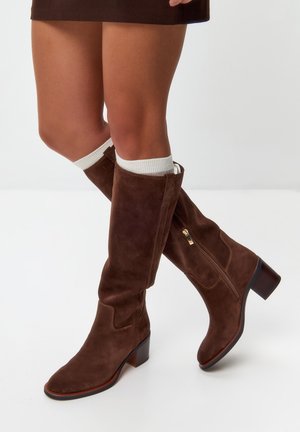 Bottes en suède marron, hauteur genou, avec un talon carré. Elles possèdent une fermeture éclair sur le côté et une texture lisse, accompagnées de chaussettes blanches côtelées.