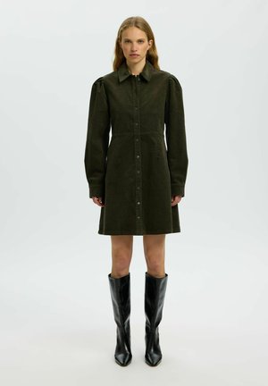 Femme portant une robe boutonnée vert foncé avec de longues manches bouffantes et un col pointu, associée à des bottes en cuir noires montant jusqu'aux genoux.