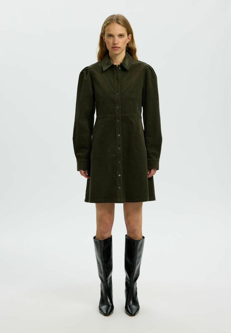 Femme portant une robe boutonnée vert foncé avec de longues manches bouffantes et un col pointu, associée à des bottes en cuir noires montant jusqu'aux genoux.