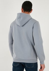 Világosszürke kapucnis pulóver, húzózsinórral, puha tapintású, bordás mandzsettákkal és alsó szegéllyel, minimális dizájn részletekkel, sötét farmerrel párosítva.