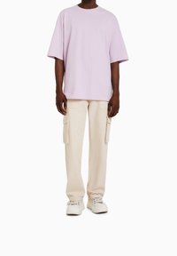 Levandulové bavlněné tričko s krátkými rukávy, které je kombinováno s béžovými cargo kalhotami s bočními kapsami a uvolněným střihem. Na nohou jsou bílé tenisky.