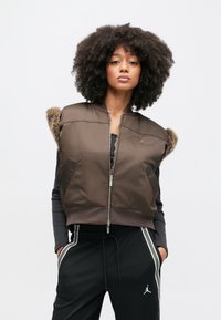 Καφέ κοντό γιλέκο με γούνινες λεπτομέρειες στους ώμους, κλείσιμο με φερμουάρ και το λογότυπο της Jordan. Φορεμένο πάνω από μαύρο μπλουζάκι με μακριά μανίκια και συνδυασμένο με μαύρο παντελόνι.