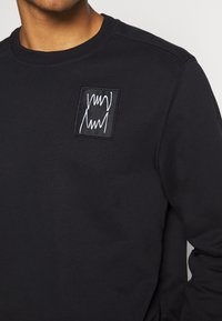 Czarna bluza z bawełny, z okrągłym dekoltem, długimi rękawami i prostokątną naszywką z białymi abstrakcyjnymi liniami na piersi.