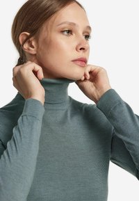 Eine Nahaufnahme eines Rollkragenpullovers in einem gedämpften blau-grauen Farbton, mit strukturiertem Rippdesign und enganliegender Silhouette. Das Model richtet den Kragen.