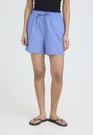 Person iført lyseblå shorts med snor, sort tanktop, sorte klipklappere, stående mod en ensfarvet lys baggrund.