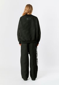 Sudadera negra texturizada con cuello redondo y un parche en la parte posterior, combinada con pantalones anchos negros que presentan una franja gráfica lateral.